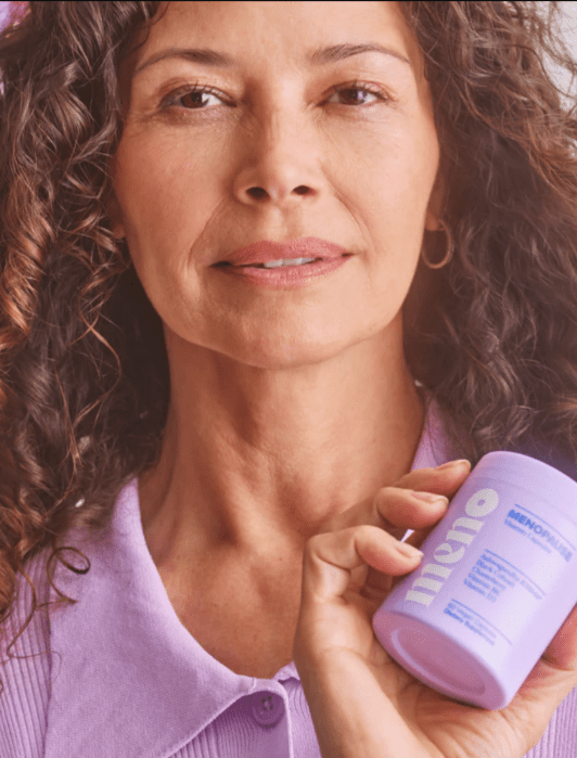 A person with curly hair wearing a lavender shirt holds a purple container of Olly's "MenoGlow" vitamins in their hand. Flowers are visible in the background.