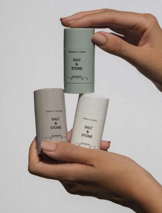 Three Salt & Stone deodorant sticks in different scents are held by hands against a plain background.