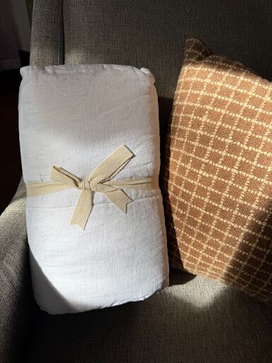A rolled white comforter tied with a beige ribbon rests on a gray chair next to a brown patterned pillow.