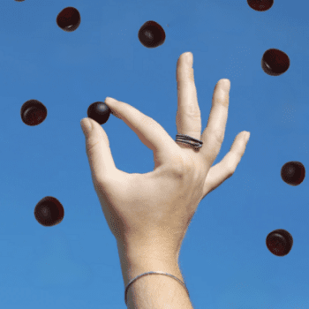 A hand with rings holds a small black sphere against a clear blue sky, surrounded by similar floating spheres.