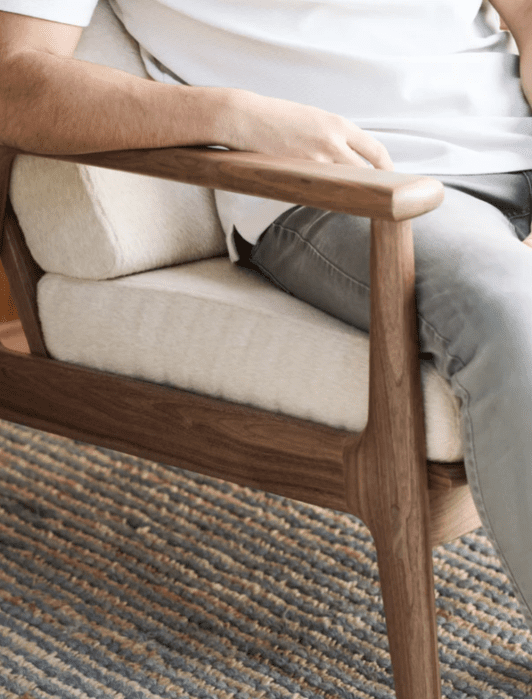 A person sitting on a wooden chair with beige cushions, wearing a white shirt and gray pants.