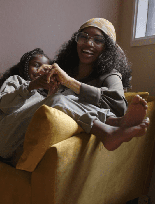 A person and a child with long curly hair laugh together while sitting on a mustard-colored armchair. A small yellow lamp and a potted plant are on a nearby table.