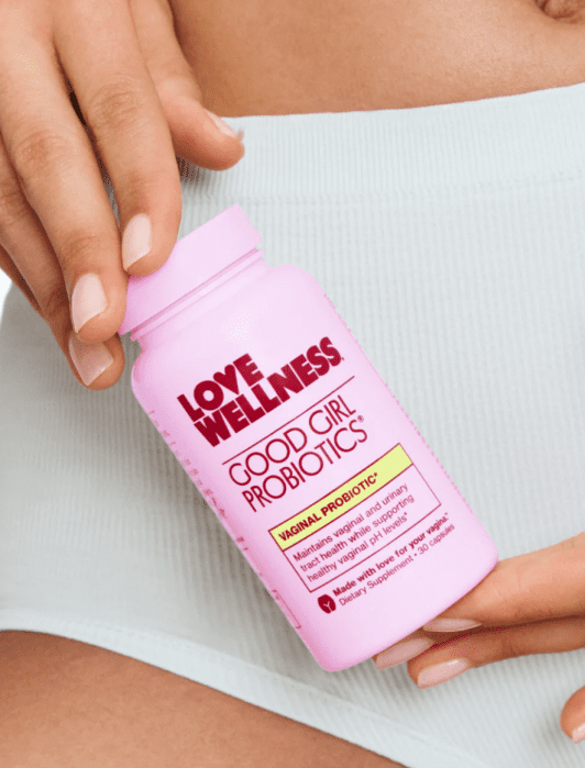 A person holding a pink bottle of "Love Wellness Good Girl Probiotics" against their waist, wearing light-colored shorts.