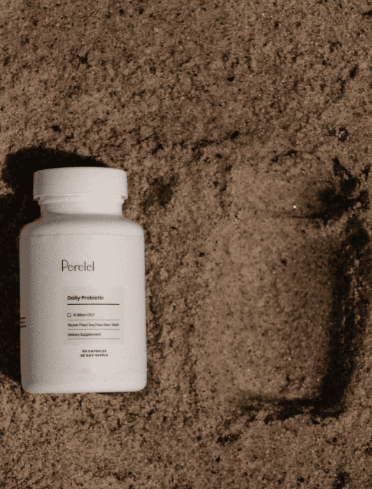 A bottle of Perelel Daily Probiotic is placed on sand next to its imprint.