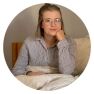 A person with glasses and a striped shirt is sitting on a bed, thoughtfully resting an arm on their chin while contemplating an upcoming mattress review.