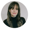 A person with long brown hair and bangs, wearing a black turtleneck and hoop earrings, smiles softly in front of a plain background, reminiscent of the serene comfort found in an organic mattress.