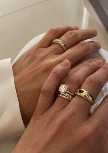 Two hands with gold rings. One hand has multiple rings, including one with a large stone, and a ring with a red stone. The other hand has a simpler band with small stones.