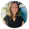 A smiling woman with long brown hair wearing a black top stands in front of a blue wall, adding charm to the serene setting. Behind her, a lush plant hints at nature's beauty—just like choosing an organic mattress for a healthier night's sleep.