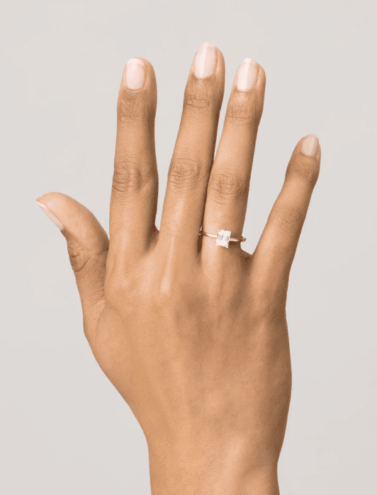 A hand wearing a gold ring with a square-cut gemstone on the ring finger, positioned against a plain background.