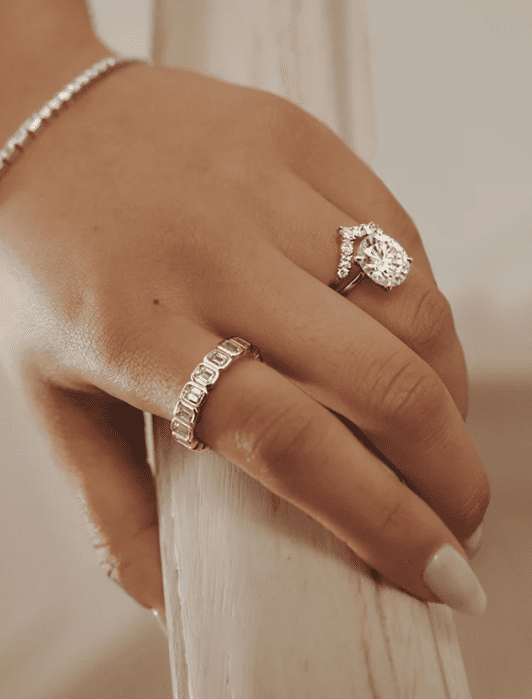 A hand wearing a diamond ring, a gold band with gems, and a silver bracelet rests on a railing.