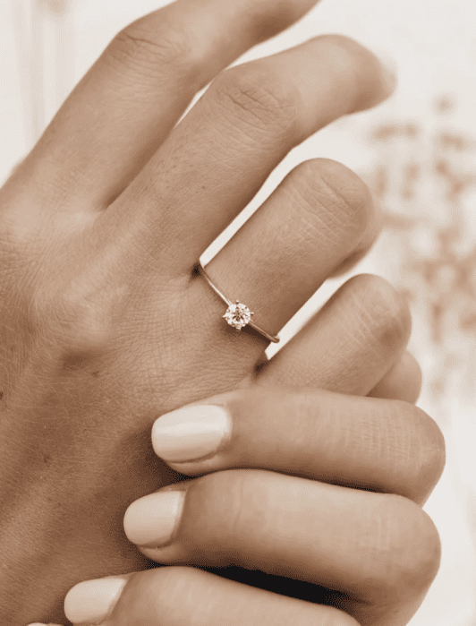 A hand wearing a simple gold ring with a single diamond is shown against a soft, neutral background.