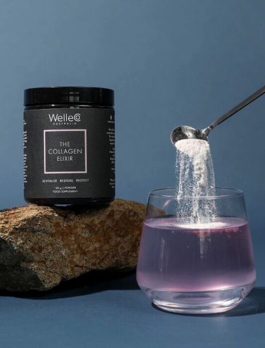 A hand pours collagen powder from a jar into a glass of pink liquid, with a rock in the background.