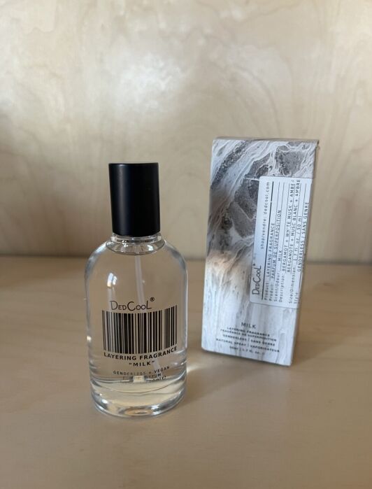 A bottle of DedCool Layering Fragrance Milk stands next to its box on a light wooden surface.