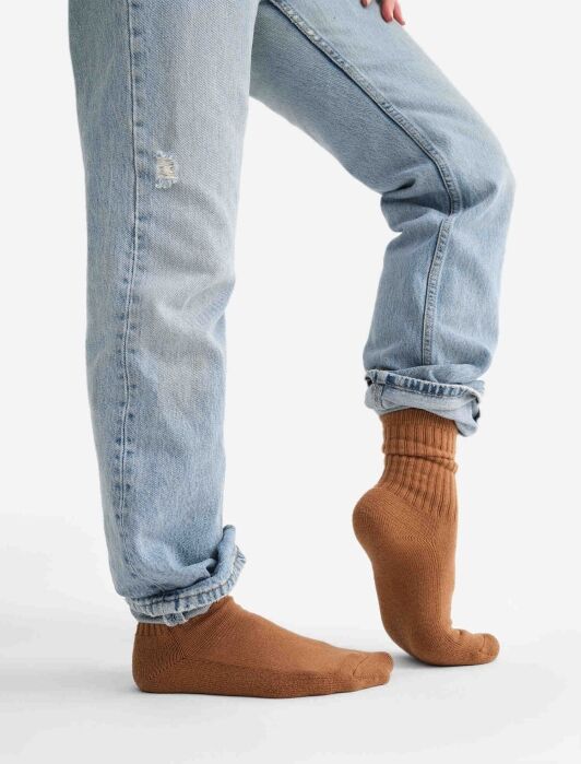 Person wearing light blue jeans and brown socks standing on one foot, with the other foot raised behind, against a plain white background.
