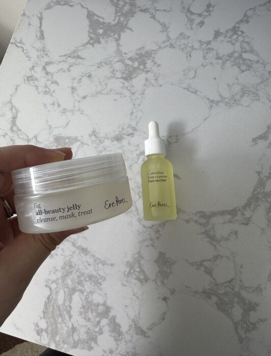 Hand holding a jar of fit all-beauty jelly next to a dropper bottle labeled Australian face nectar by Ere Perez, on a marble surface.