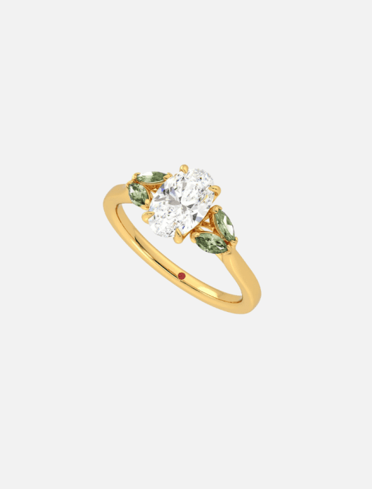 Gold ring with three stones: a central oval white gem and two green gems on either side, set on a gold band with a small red dot detail.