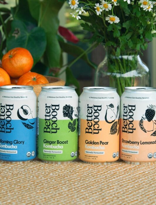 Four cans of Better Booch kombucha in different flavors on a woven surface, with a wooden bowl of oranges and green foliage in the background.