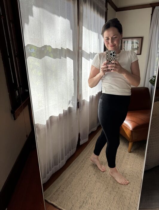 A woman stands barefoot on a rug, taking a mirror selfie in a bright room with sheer curtains and a brown chair.