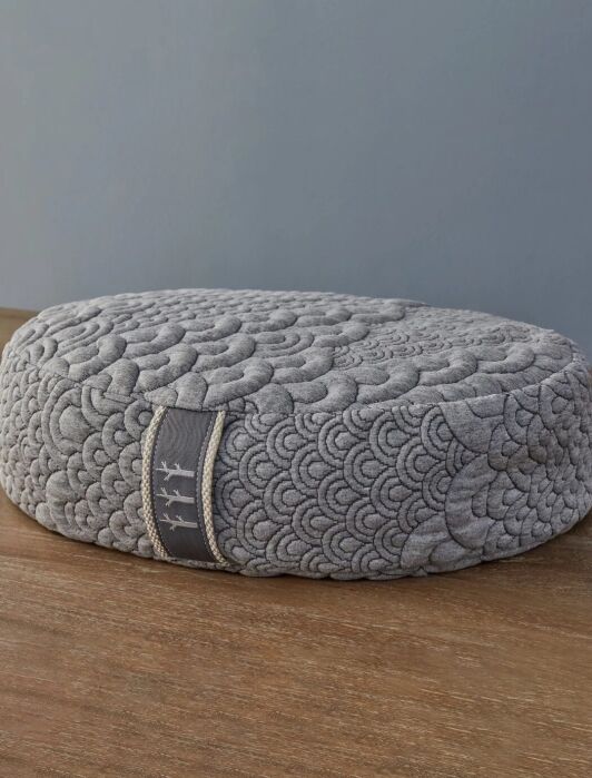 Round gray cushion with a quilted pattern, featuring a fabric loop with decorative stitching, placed on a wooden surface against a gray background.
