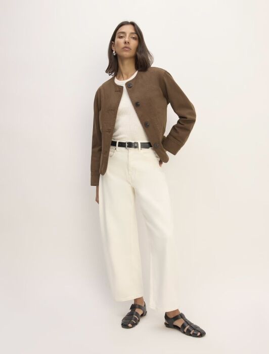 A person poses against a plain backdrop wearing a brown jacket, white top, wide-legged white pants, black belt, and black sandals.