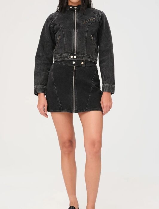 Person wearing a black denim jacket and skirt with zipper details, paired with black loafers, stands against a plain white background.