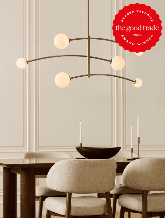Modern dining room with a brass chandelier, beige upholstered chairs, a wooden table set with candlesticks, and an award badge reading “The Good Trade 2026 Reader Favorite.”.