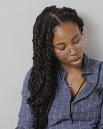 A person with long twisted hair and glasses looks down, wearing a blue checkered shirt and a necklace against a plain background.