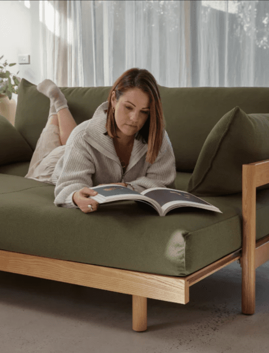 A woman is lying on a green sofa, reading a book. She is wearing a white sweater and beige pants. The room is softly lit with sheer curtains in the background.