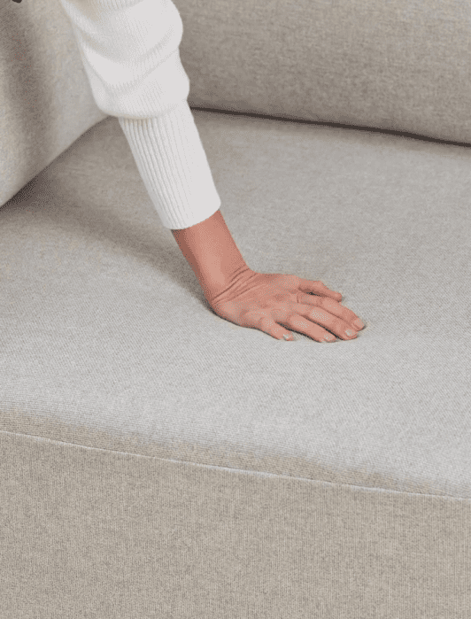 A hand presses down on a light gray couch cushion, showing the fabric and texture.