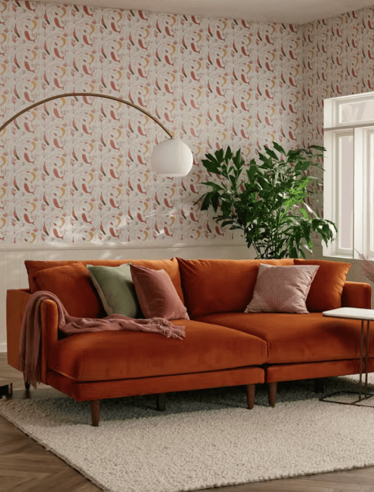 Living room with a rust-colored sofa, pillows, a white floor lamp, a potted plant, and patterned wallpaper. A small side table and large window are also visible.