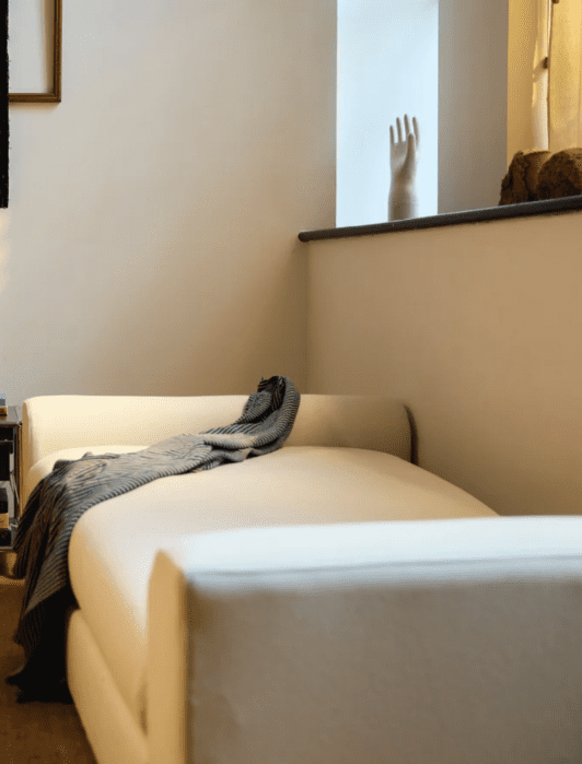 A cozy living room corner with a white sofa draped with a striped blanket, a small side table with books and decor, a window ledge with a hand sculpture, and warm lighting.