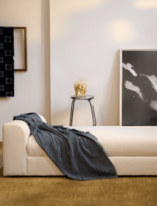 Minimalist room with a white daybed, striped blanket, stool with decorative item, framed photographs on walls, and green carpet.