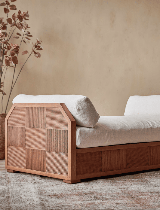 Wooden daybed with white cushions on an ornate rug, surrounded by plants against a beige wall.