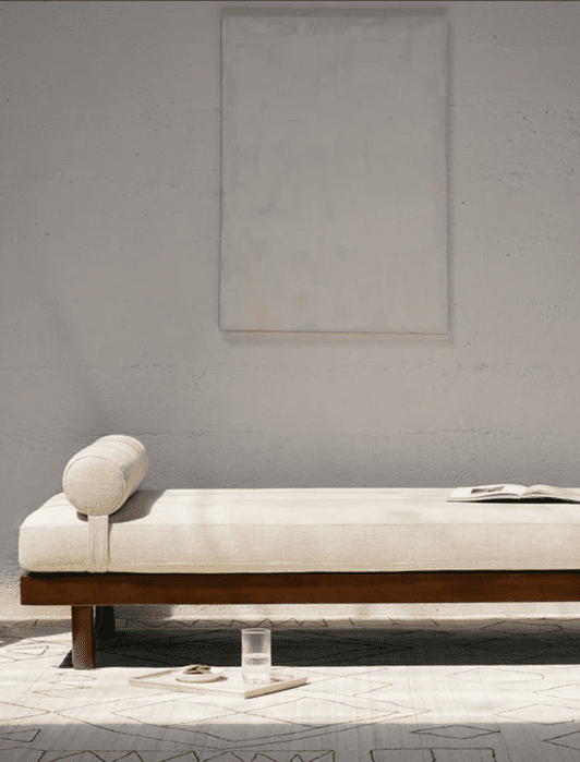 Minimalist room with a wooden daybed featuring a cylindrical pillow, open book, and a tray with a glass of water and a snack. A blank canvas hangs on the wall behind.