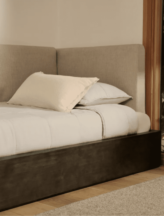A neatly made bed with a beige headboard is positioned next to a window showing green plants outside.