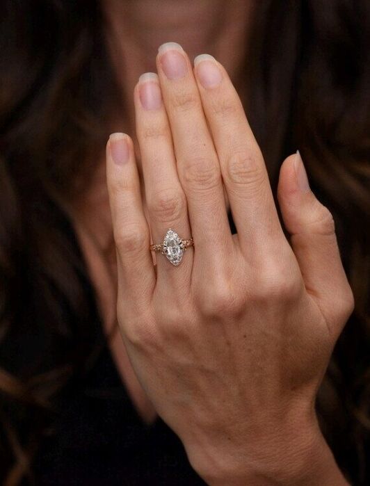 A person with long brown hair shows a hand with a diamond ring on the ring finger against a dark background.