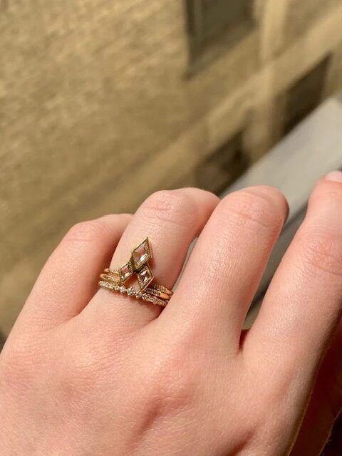 A hand with a stack of rings, including a large marquise-shaped one, in natural light near a window with a brick wall backdrop.