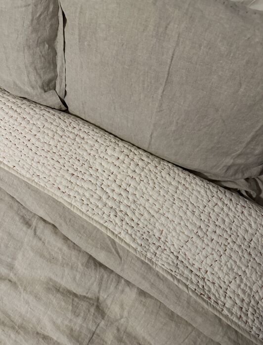 A neatly made bed with beige linen sheets, a matching pillow, and a textured white quilted blanket folded beneath the pillow.
