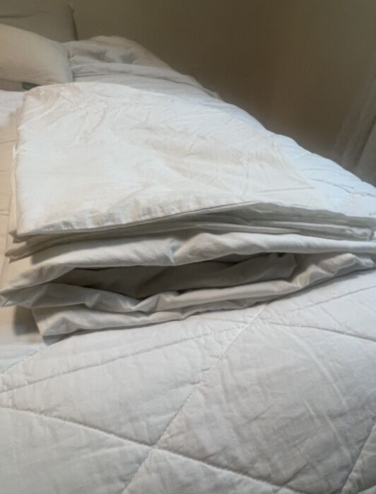 A neatly folded white blanket lays on a white quilted bedspread in a dimly lit room.