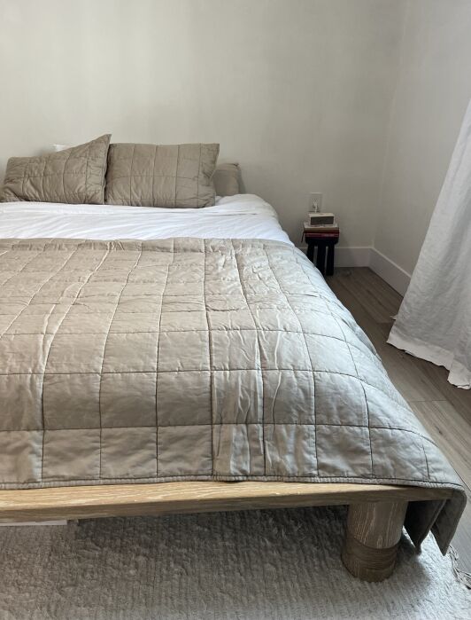 A neatly made bed with a beige quilt, matching pillows, and wooden bed frame. A small nightstand with a book and lamp is visible next to the bed. A white curtain hangs on the right.