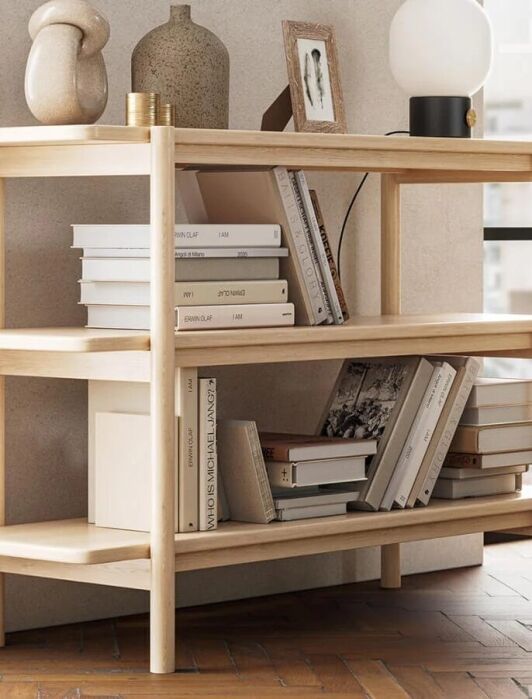 Wooden bookshelf with three shelves, holding various books, vases, and a lamp. Positioned near a window with a green sofa nearby.