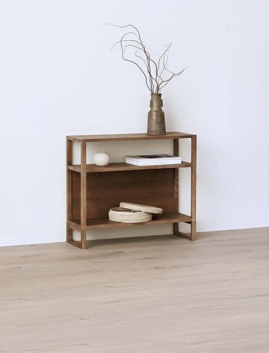 A wooden shelf against a white wall holds a book, a small vase with branches, a round white object, and two woven baskets on the middle shelf.