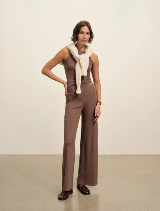 A woman stands indoors wearing a sleeveless brown knit jumpsuit, a cream sweater draped over her shoulders, and brown shoes.