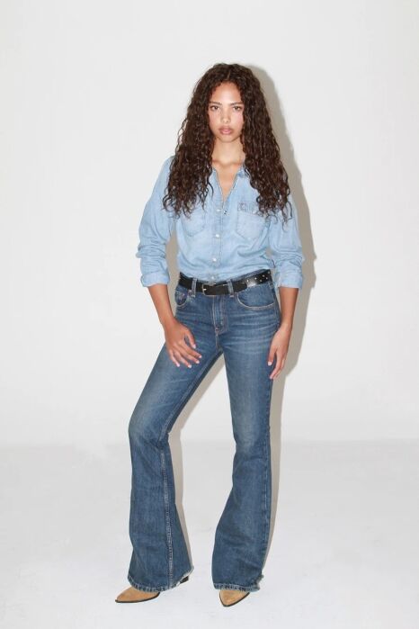 A woman with long curly hair wears a light blue denim shirt tucked into high-waisted blue jeans with a black belt and tan pointed shoes, standing against a plain white background.