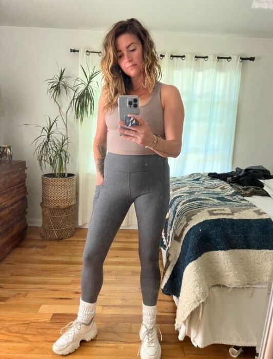 A woman stands in front of a mirror taking a selfie. She is wearing a tank top, gray leggings, white socks, and sneakers. A bed and plants are visible in the background.