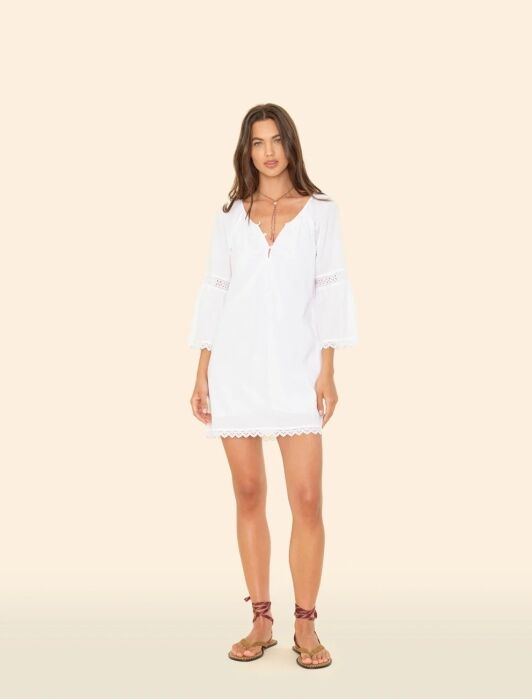 Woman standing against a beige background, wearing a white long-sleeve, V-neck dress with lace trim, brown sandals, and layered necklaces.