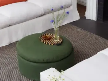 A round green ottoman with a checkered tray, glass vase with blue flowers, and a small bowl sits on a patterned rug between two white sofas.
