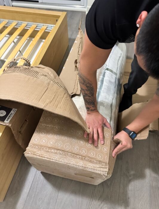 A person unboxes a rolled-up mattress from a branded cardboard box next to a partially assembled wooden bed frame.