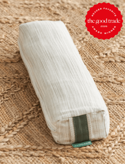 A rectangular, off-white fabric pillow with a green tag labeled "Avocado" rests on a woven rug. A red badge in the corner reads "Reader Favorite The Good Trade 2026 Award Winner.