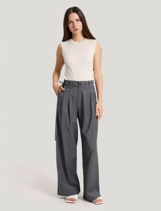 A woman stands against a plain background wearing a sleeveless white top, high-waisted gray wide-leg trousers, and white sandals.